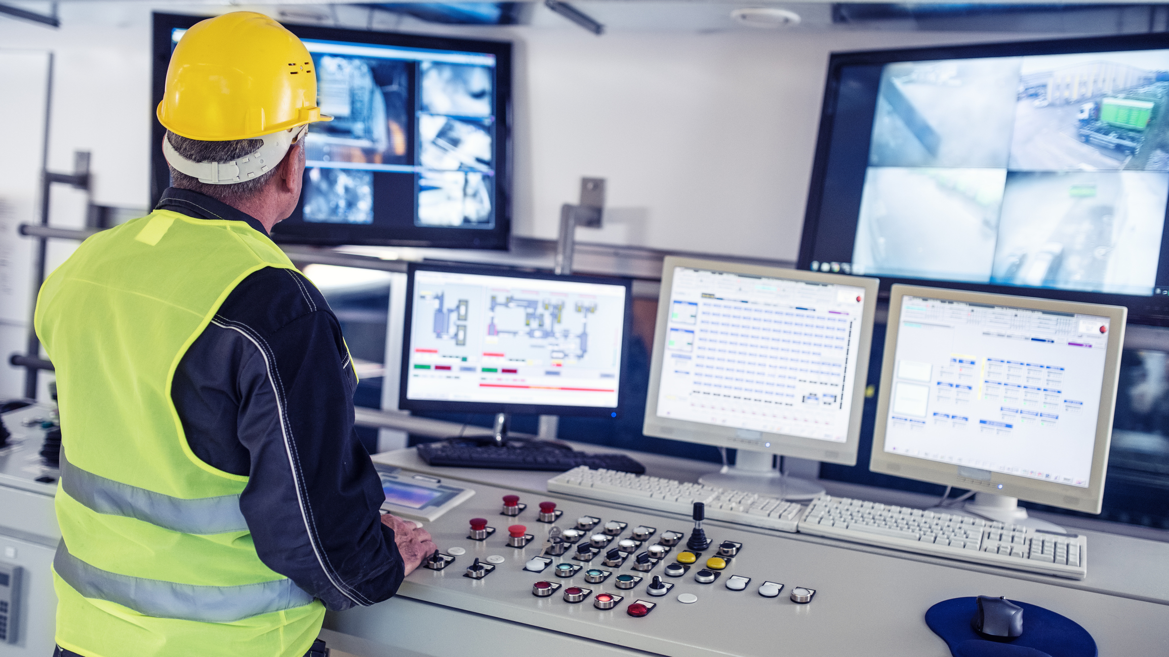 Man wearing a yellow hard hat and reflective vest operating an industrial control panel with multiple buttons, joysticks, and five computer monitors displaying technical diagrams and security camera footage.