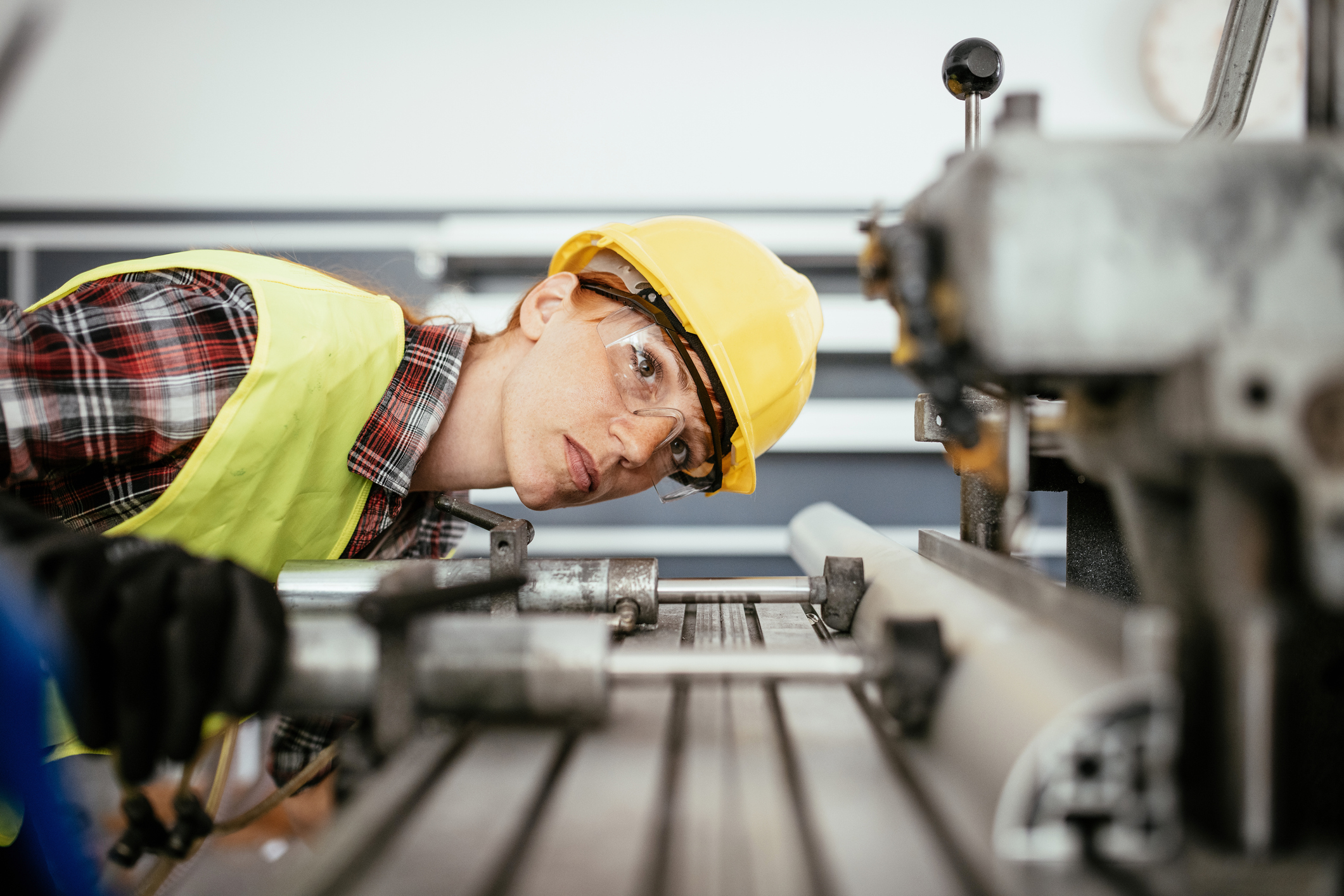 Female factory worker in a yellow safety vest, hard hat, and protective glasses closely inspecting a metal machine part on an industrial production line.