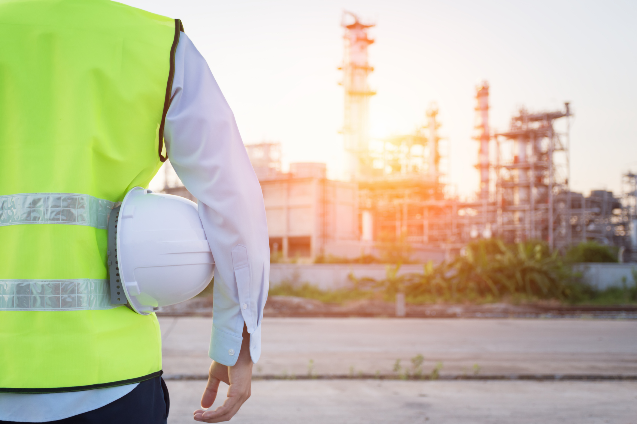 Person wearing a high-visibility safety vest and holding a white hard hat, standing outdoors near an industrial chemical plant at sunset.