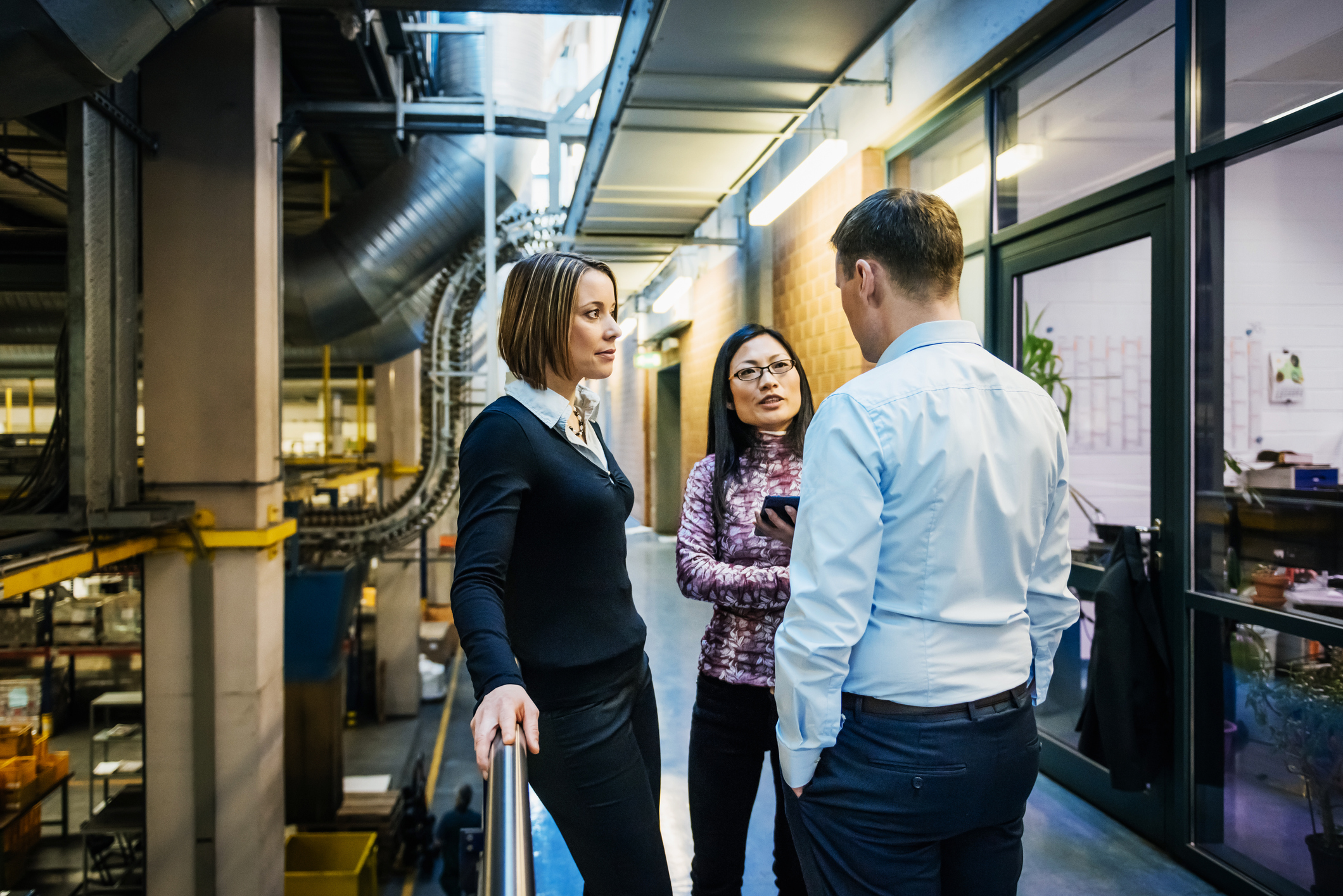 Three business professionals standing in an industrial hallway having a discussion; one woman with short hair leans on a railing, another woman wearing glasses holds a smartphone, and a man with hands in his pockets listens.