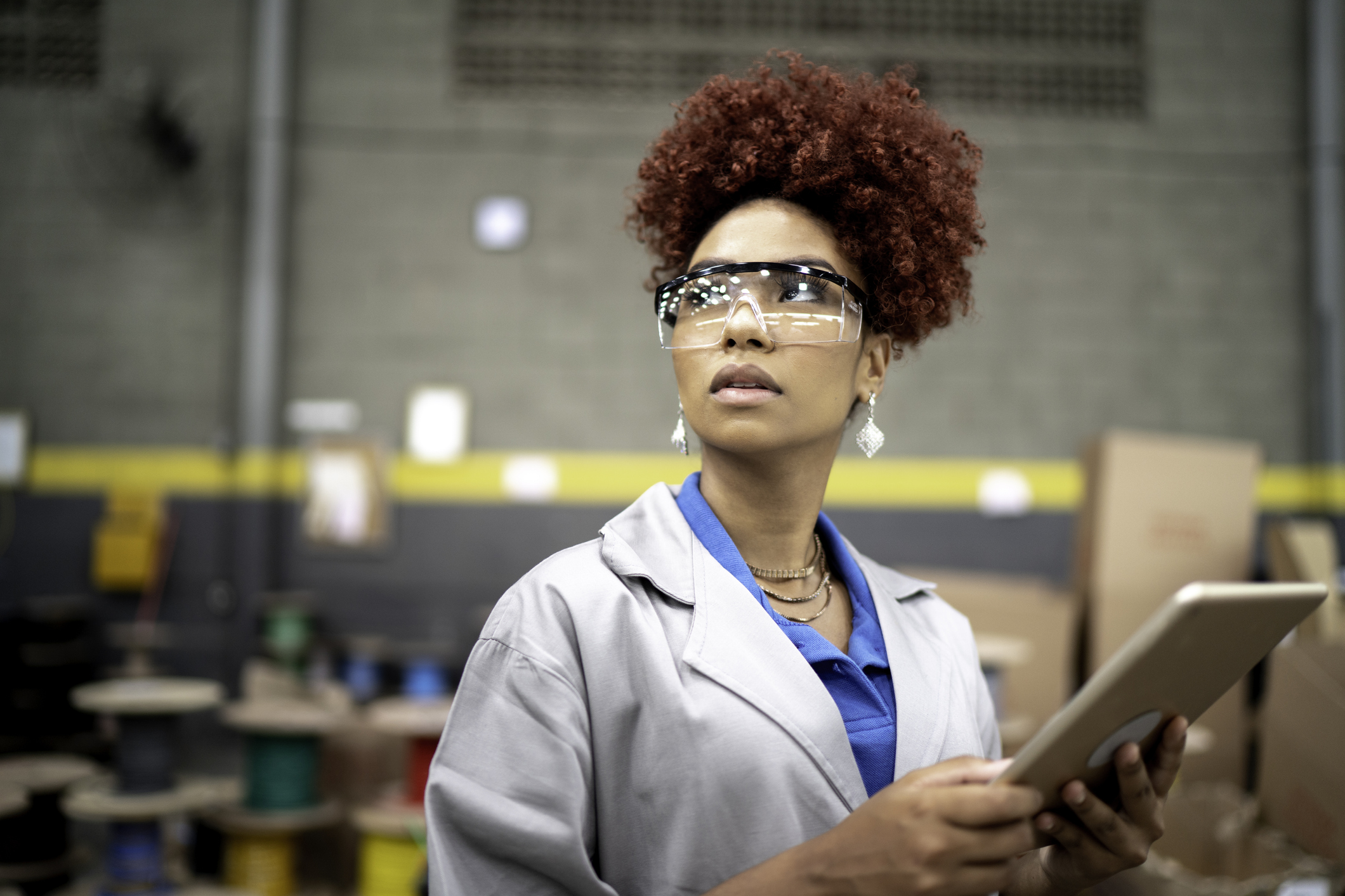 A woman with curly red hair wearing protective safety glasses, blue polo shirt, and gray lab coat holds a tablet and looks to the side in an industrial warehouse setting with spools of cable and cardboard boxes in the background.