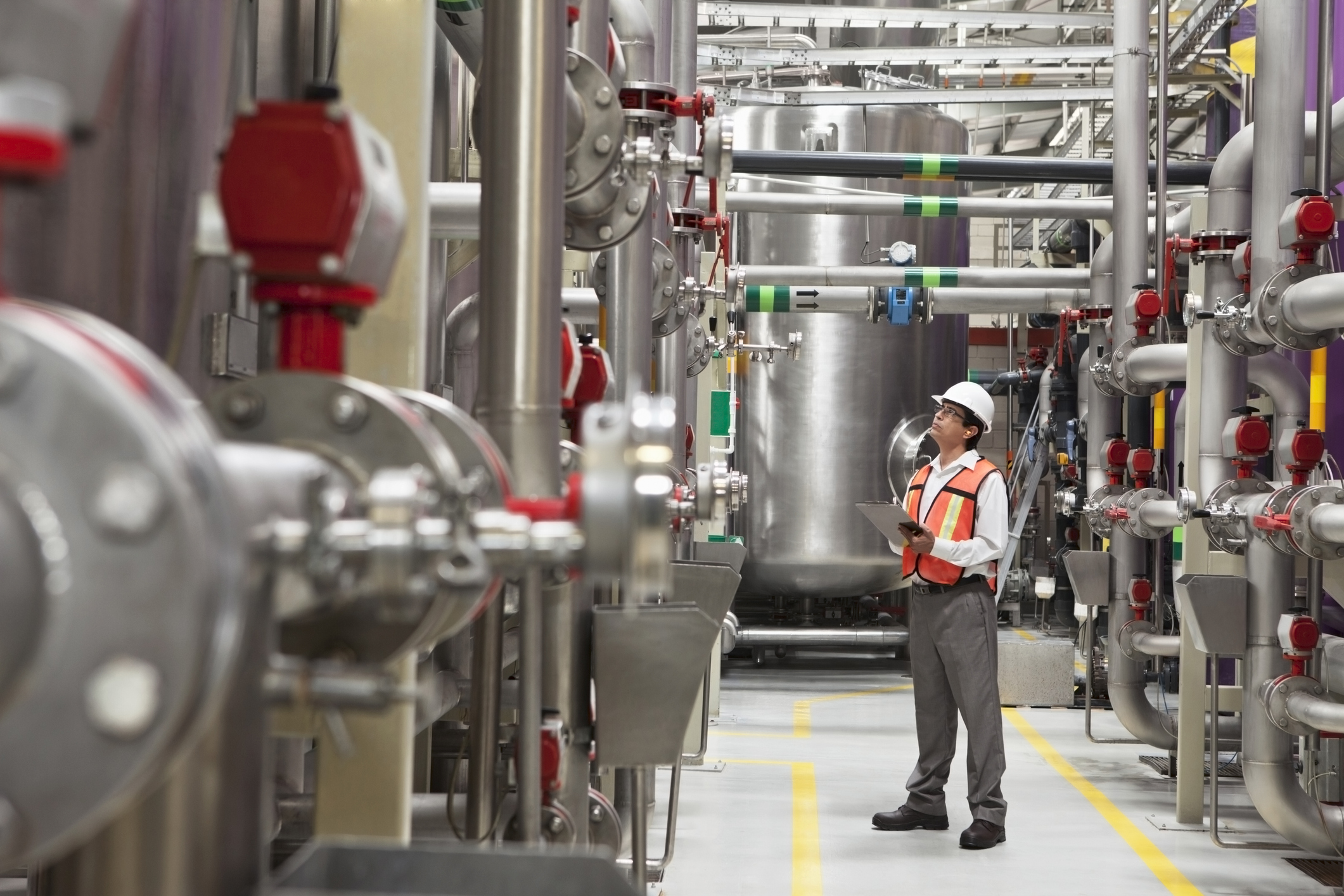 Engineer wearing a white hard hat and orange safety vest inspecting large industrial pipes and tanks inside a clean factory or plant, holding a clipboard and looking upwards attentively.