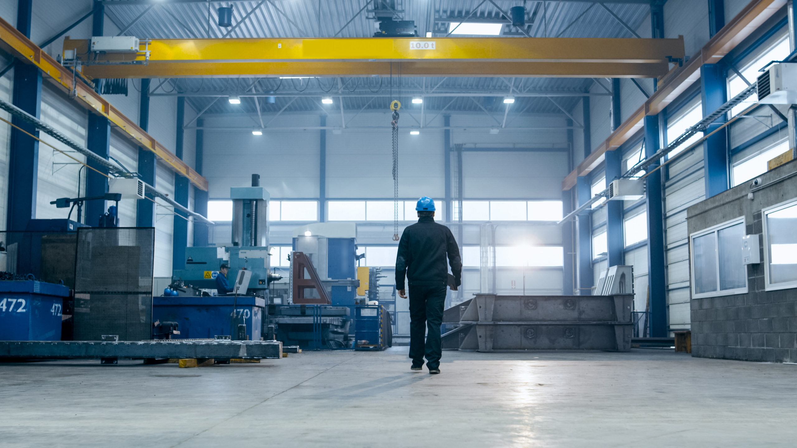 Man wearing a blue hard hat and dark work clothes walking in a spacious industrial factory with large machinery, a yellow overhead crane, and high windows letting in natural light.
