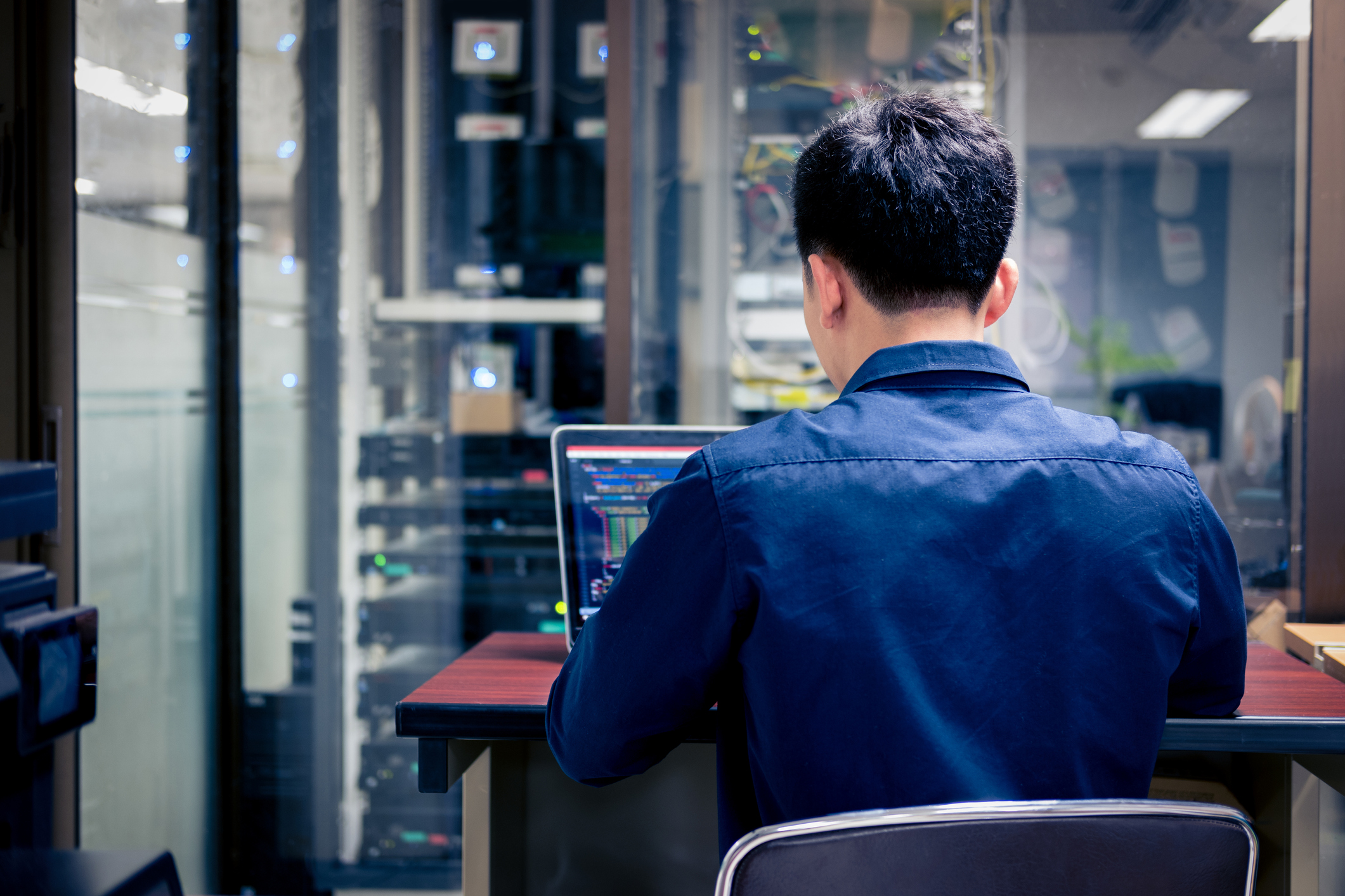 Man wearing a blue shirt working on a laptop in a server room with computer equipment racks and networking cables visible through glass panels.