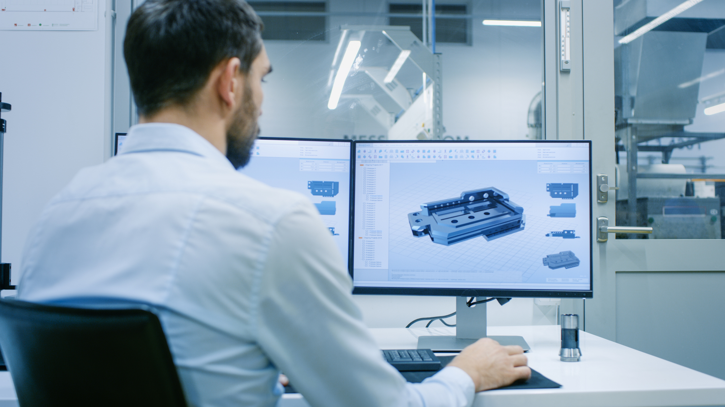 Man in a light blue shirt works on a dual-monitor setup displaying a 3D CAD model of a mechanical part in an office with glass walls and industrial equipment visible through the door.