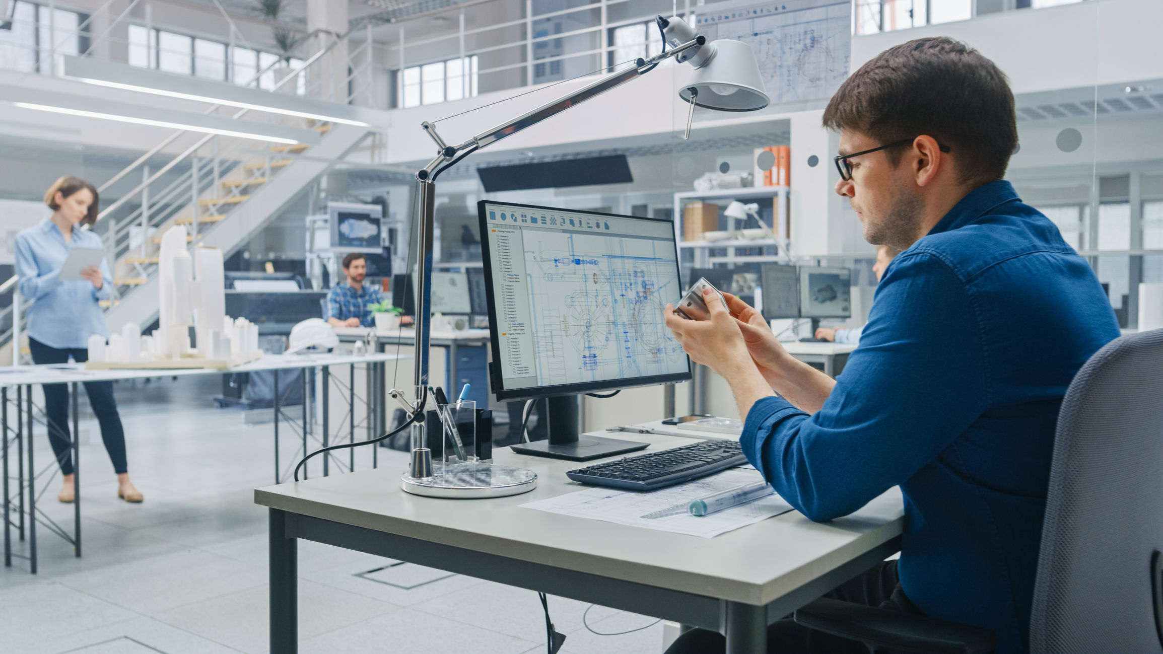 Man in glasses and blue shirt examining a small mechanical part at a desk with a computer screen displaying technical blueprints in a modern engineering office with two other people and architectural models in the background.
