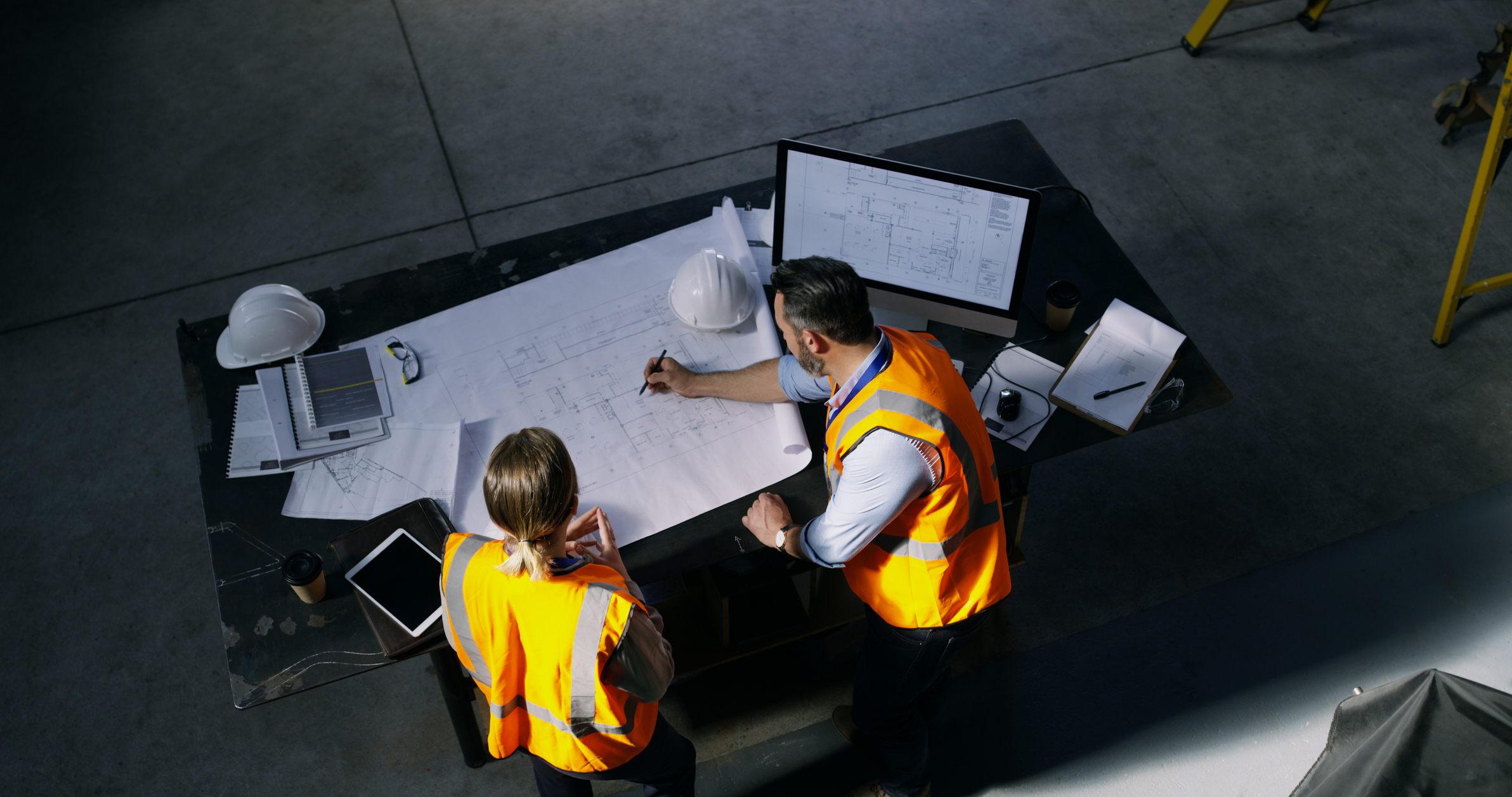 Two construction workers wearing orange safety vests reviewing blueprints on a table in a workshop; one is writing on the blueprint while the other observes; the table has a computer monitor displaying architectural plans, hard hats, documents, a tablet, and coffee cups.