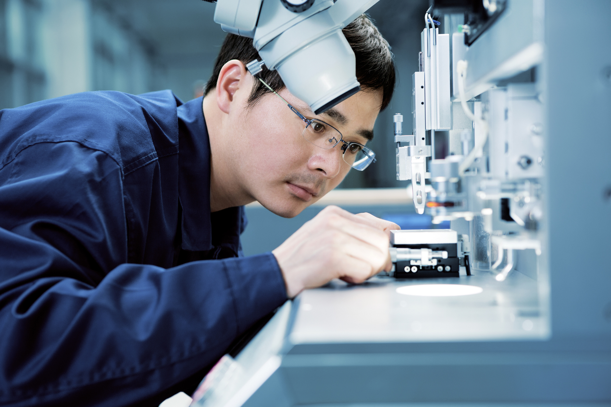 Male technician wearing glasses and a dark blue lab coat closely inspecting and adjusting a component inside a high-tech industrial machine under a microscope.