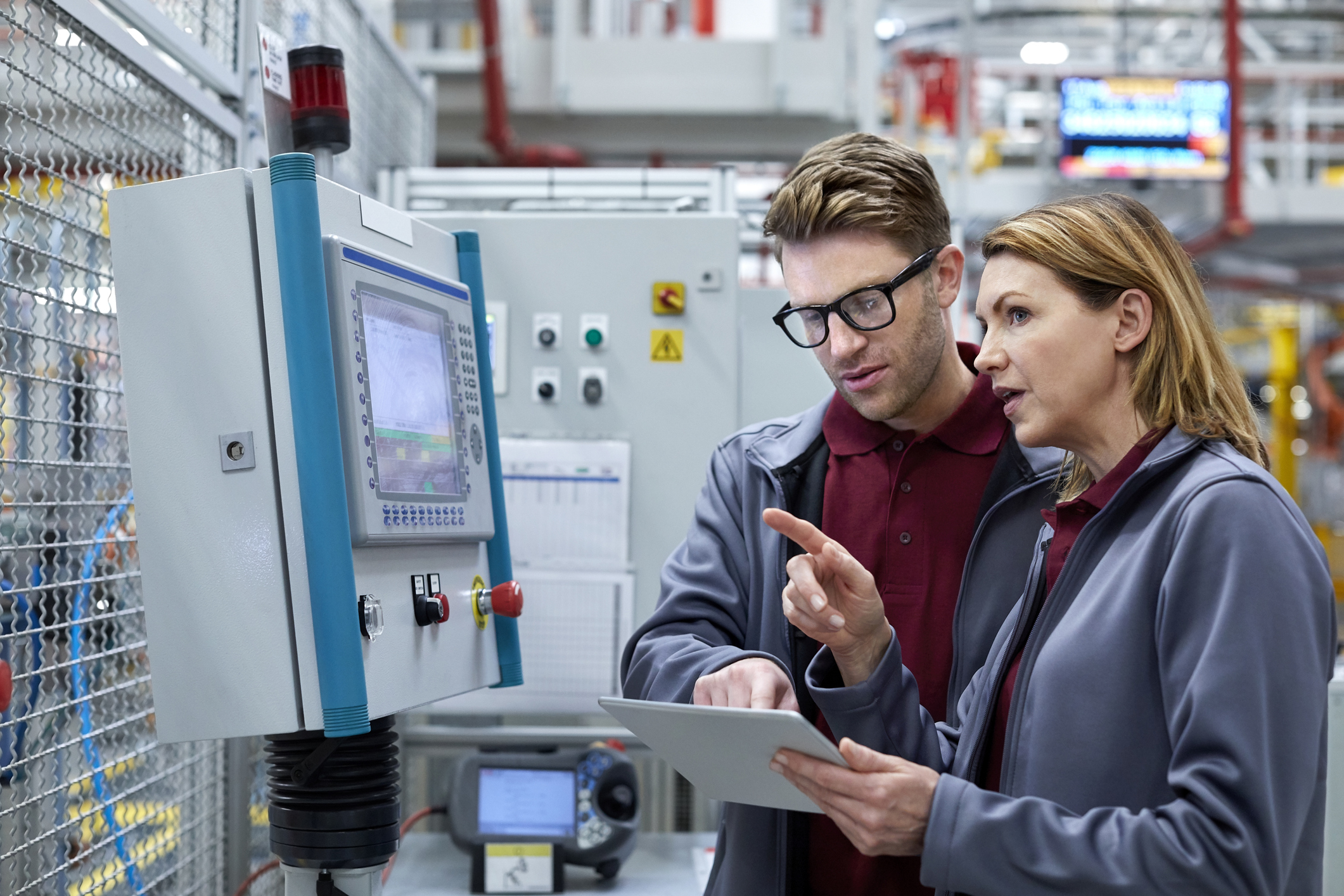 Two factory workers wearing matching gray jackets and maroon shirts, one man with glasses and one woman, discussing and pointing at a tablet in front of industrial control panels in a manufacturing setting.