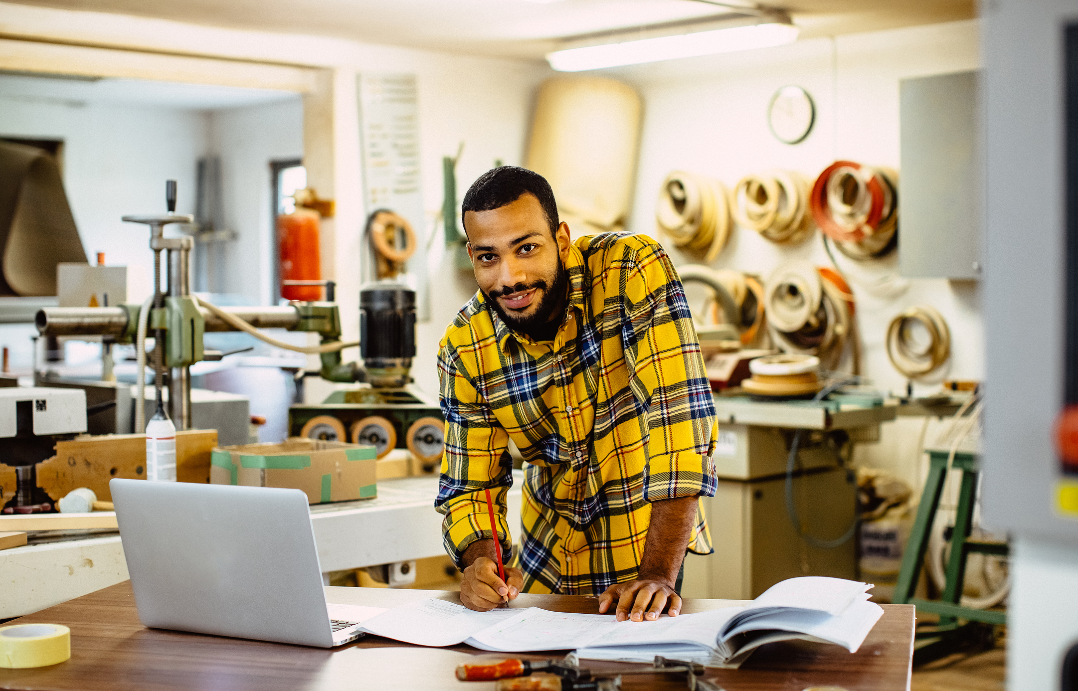 Man in a yellow plaid shirt working at a wooden table with open documents and a laptop, in a workshop filled with tools and coiled materials on the wall.