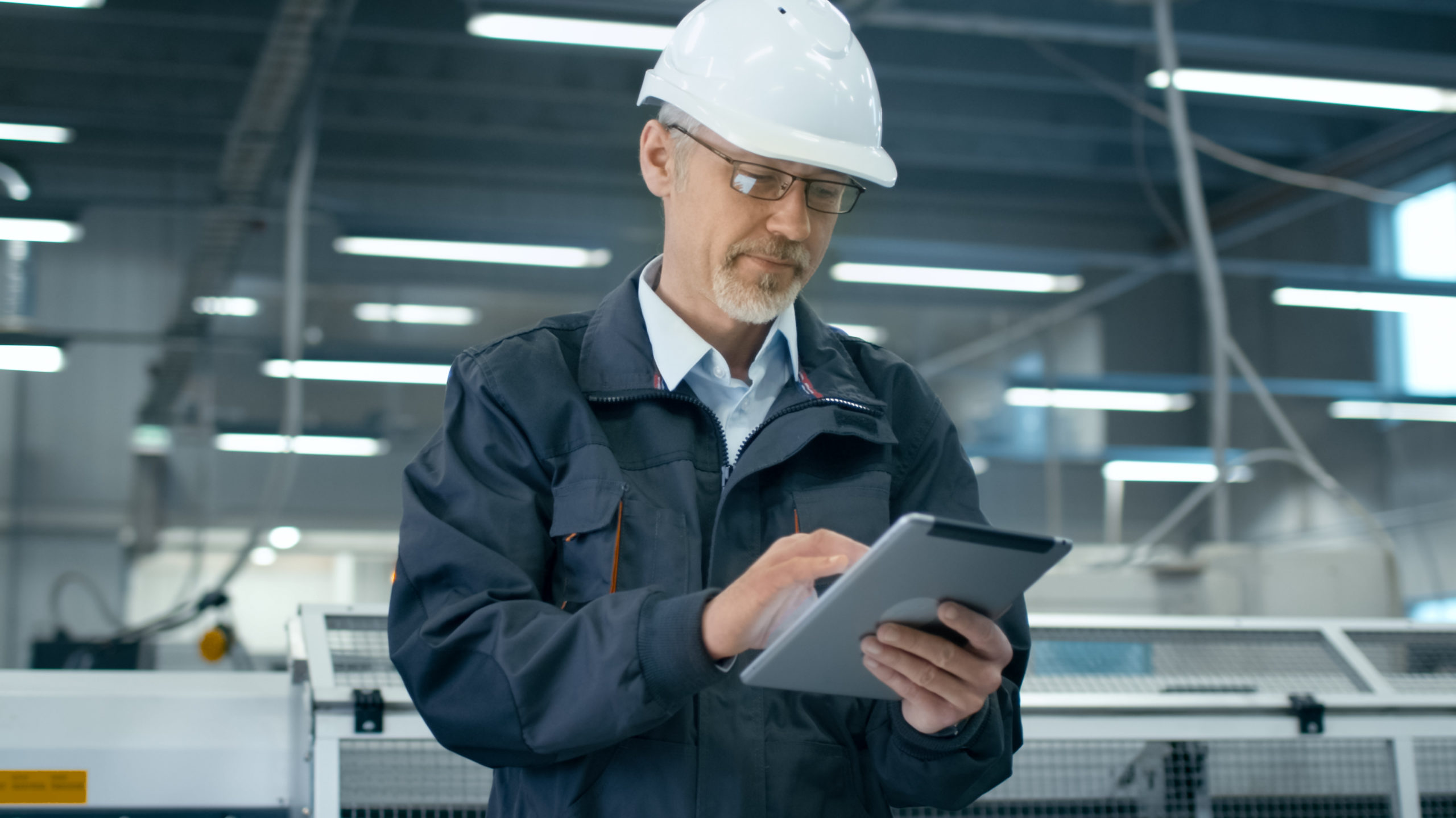 Man wearing a white hard hat and dark jacket in an industrial facility using a digital tablet.