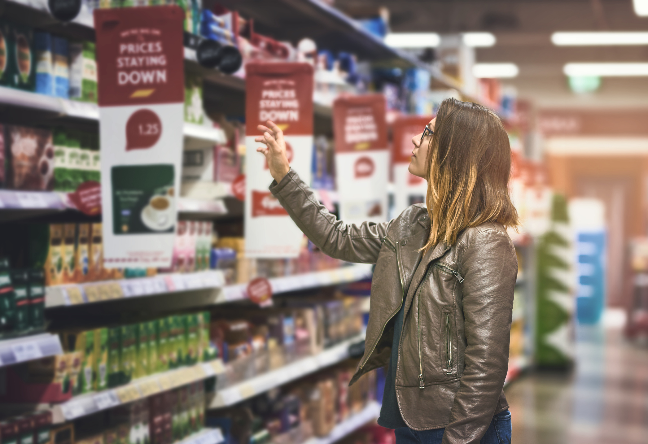 A woman wearing glasses and a brown leather jacket reaches for products on a grocery store shelf with signs indicating prices are staying down.