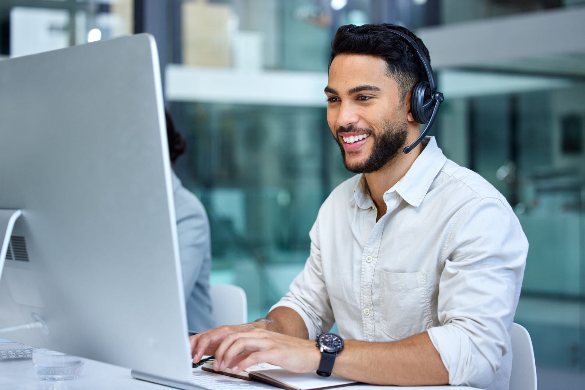 Smiling man with dark hair and beard wearing a headset, typing on a keyboard in front of a large computer monitor in a modern office.