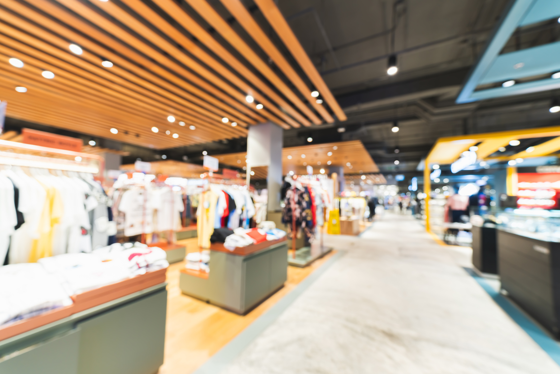 Blurred interior of a modern retail clothing store with racks of colorful clothes, wooden ceiling panels, and bright lighting.
