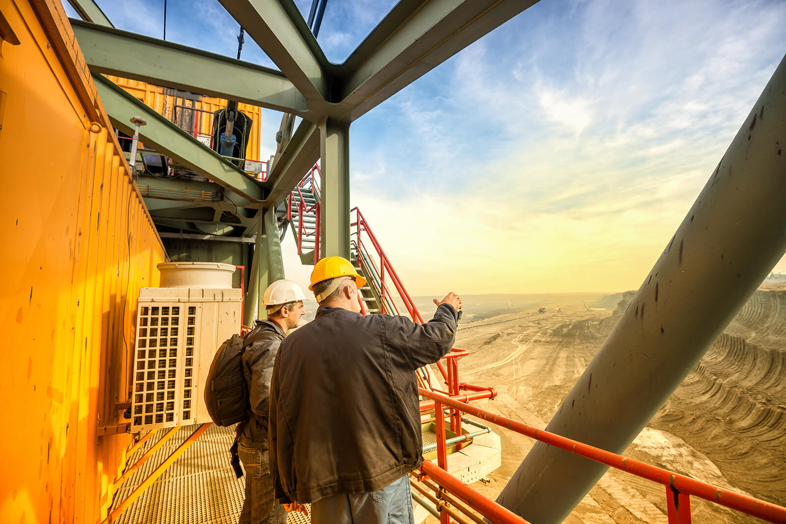 Two construction workers wearing hard hats standing on a metal platform with orange railings, overlooking a large open mining or excavation site under a partly cloudy sky, with one worker pointing towards the landscape.