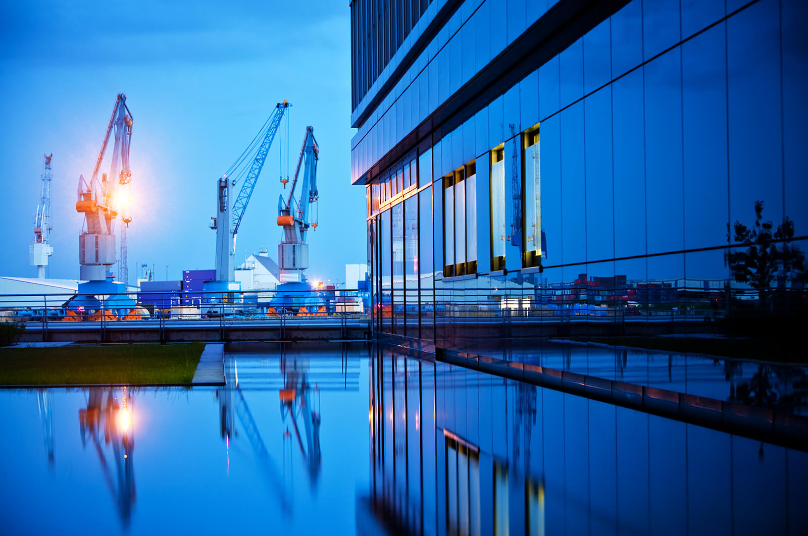 Industrial port scene at dusk with large cranes reflected in water beside a modern glass building reflecting the blue sky and lights.