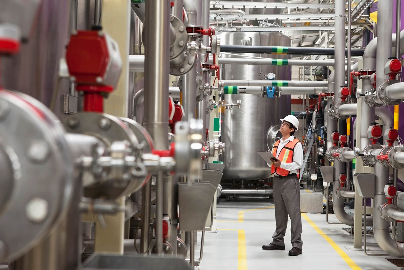 Engineer in a white hard hat and orange safety vest holding a clipboard, inspecting large stainless steel industrial pipes and tanks in a factory setting.