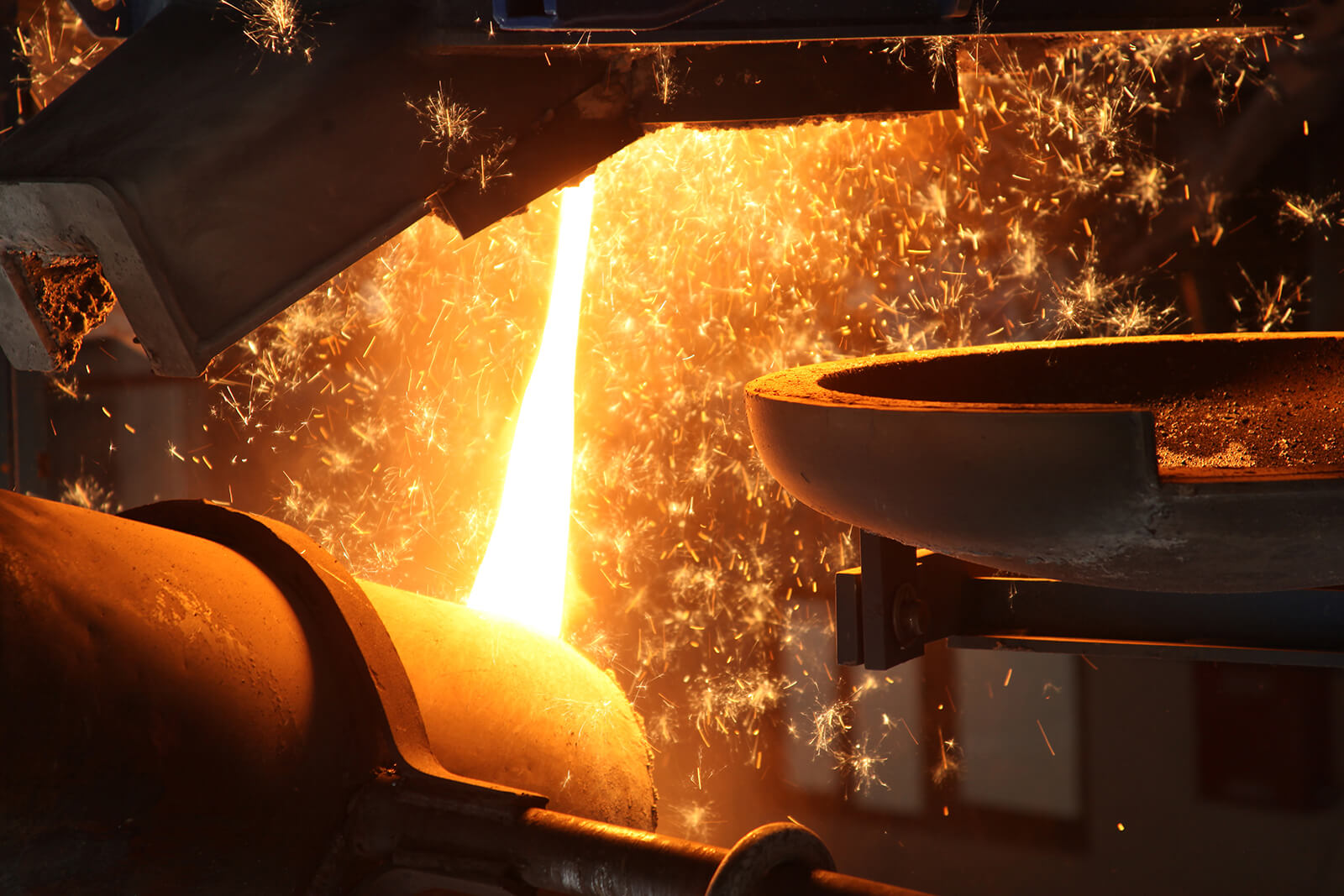 Molten metal being poured from a furnace into a container with sparks flying in an industrial foundry setting.