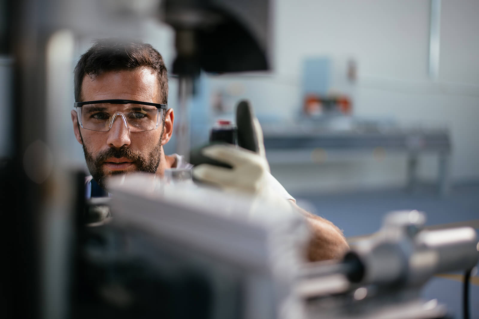 Male industrial worker wearing safety glasses and gloves closely inspecting a piece of machinery in a workshop.