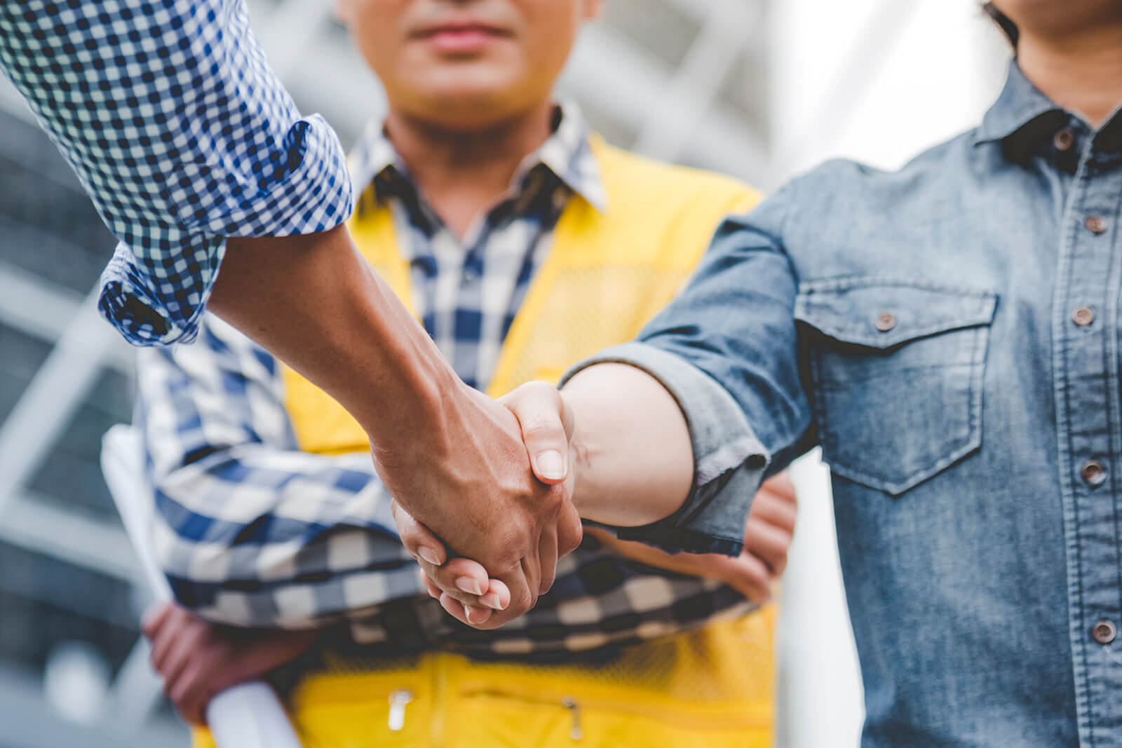 Close-up of a handshake between two people, one wearing a blue checkered shirt with rolled-up sleeves and the other with a blue denim shirt, while a person in a yellow safety vest stands in the background with arms crossed.