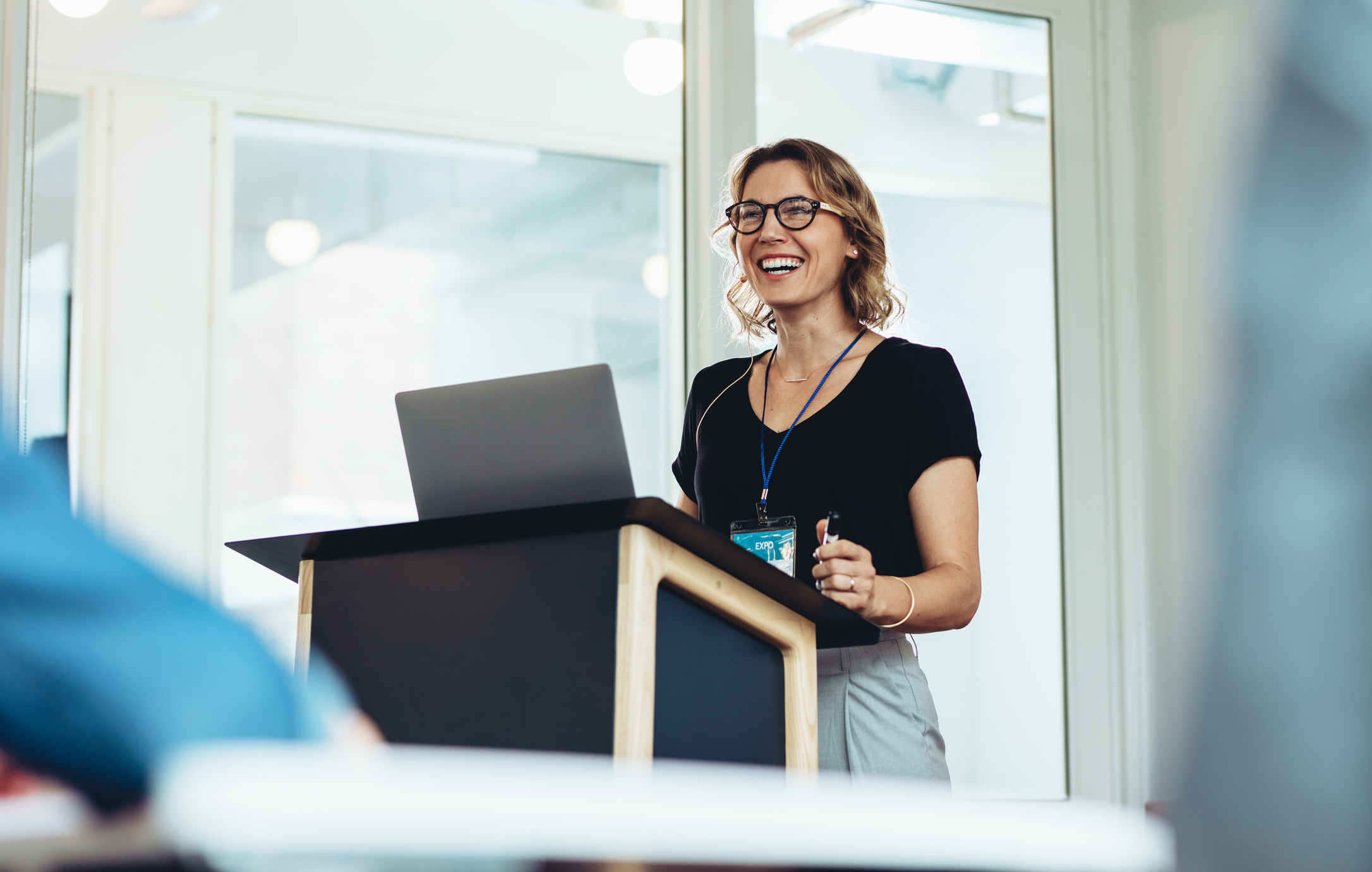 A woman with glasses and a name badge smiling while standing and speaking behind a podium with a laptop in an indoor, well-lit office setting.