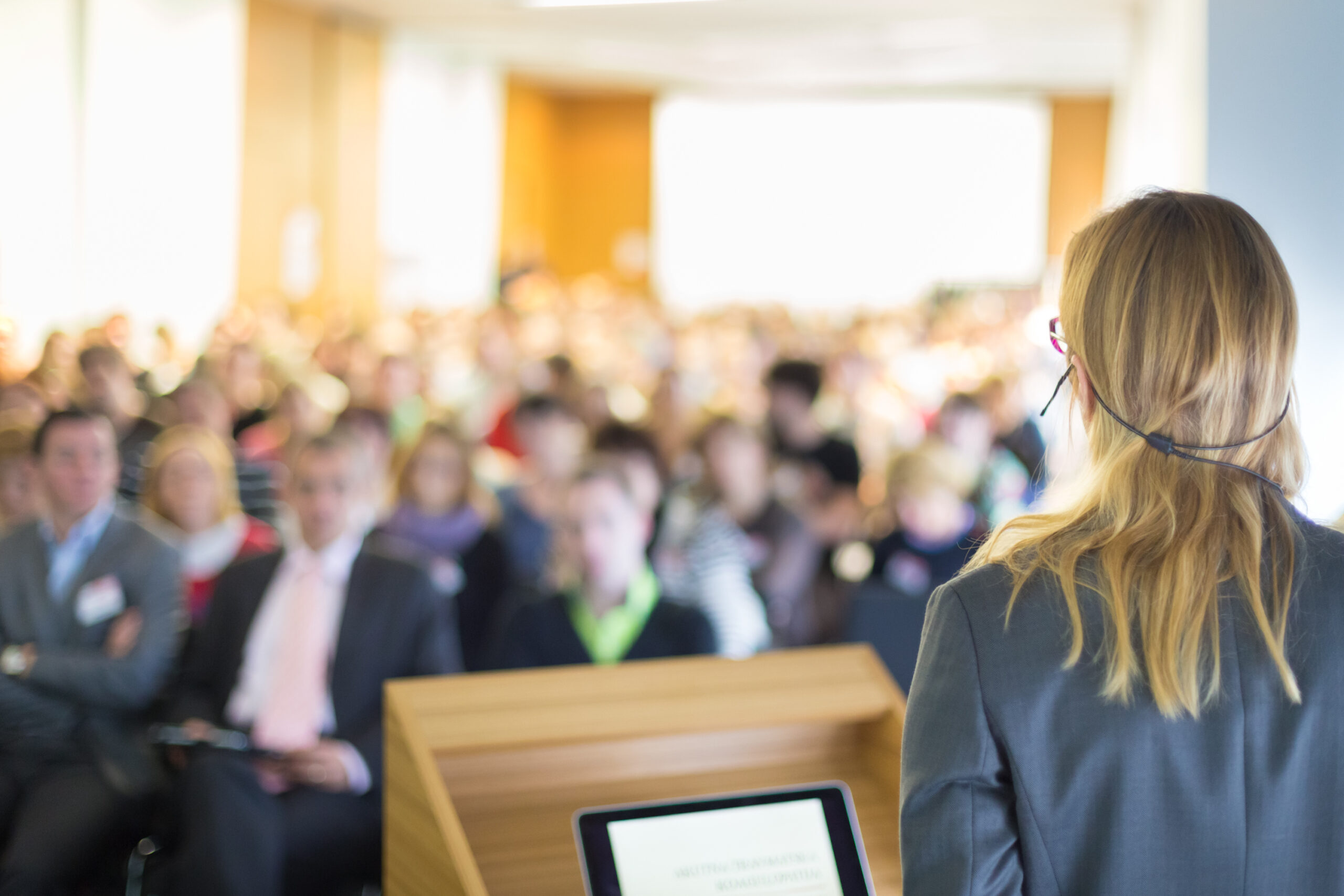 A woman with long blonde hair wearing glasses and a headset microphone speaks at a podium to a large, blurred audience in a bright room.