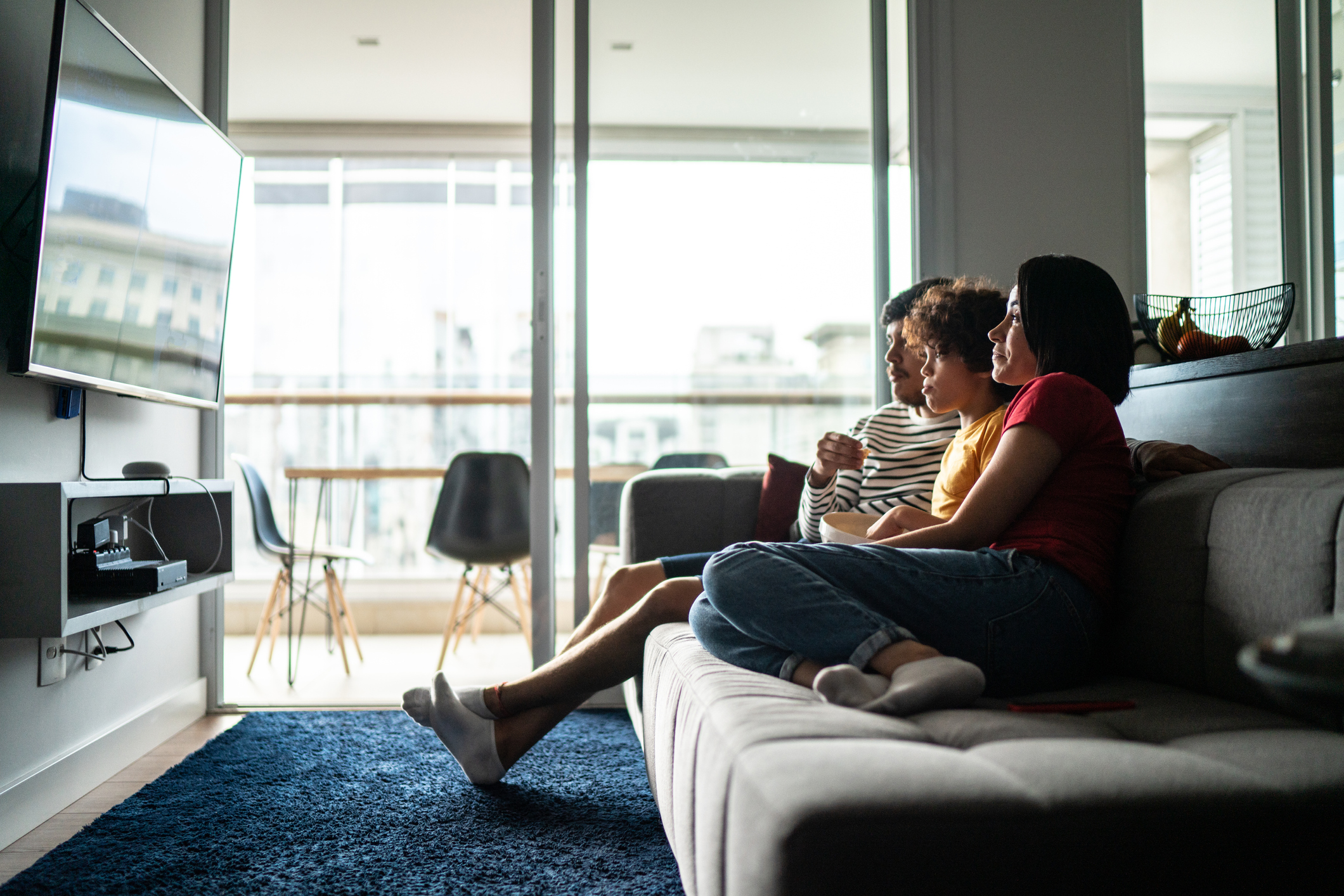 A family of three sitting closely on a gray couch in a living room, watching a wall-mounted TV with a large window and balcony in the background.