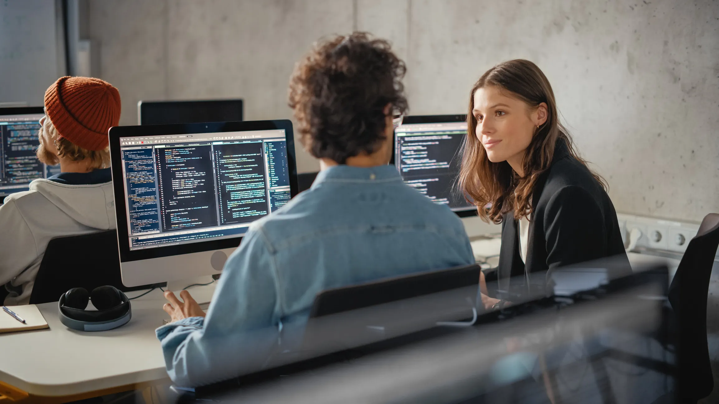 Two people sitting at desks in an office with computer screens displaying code, one person explaining or discussing something with the other.