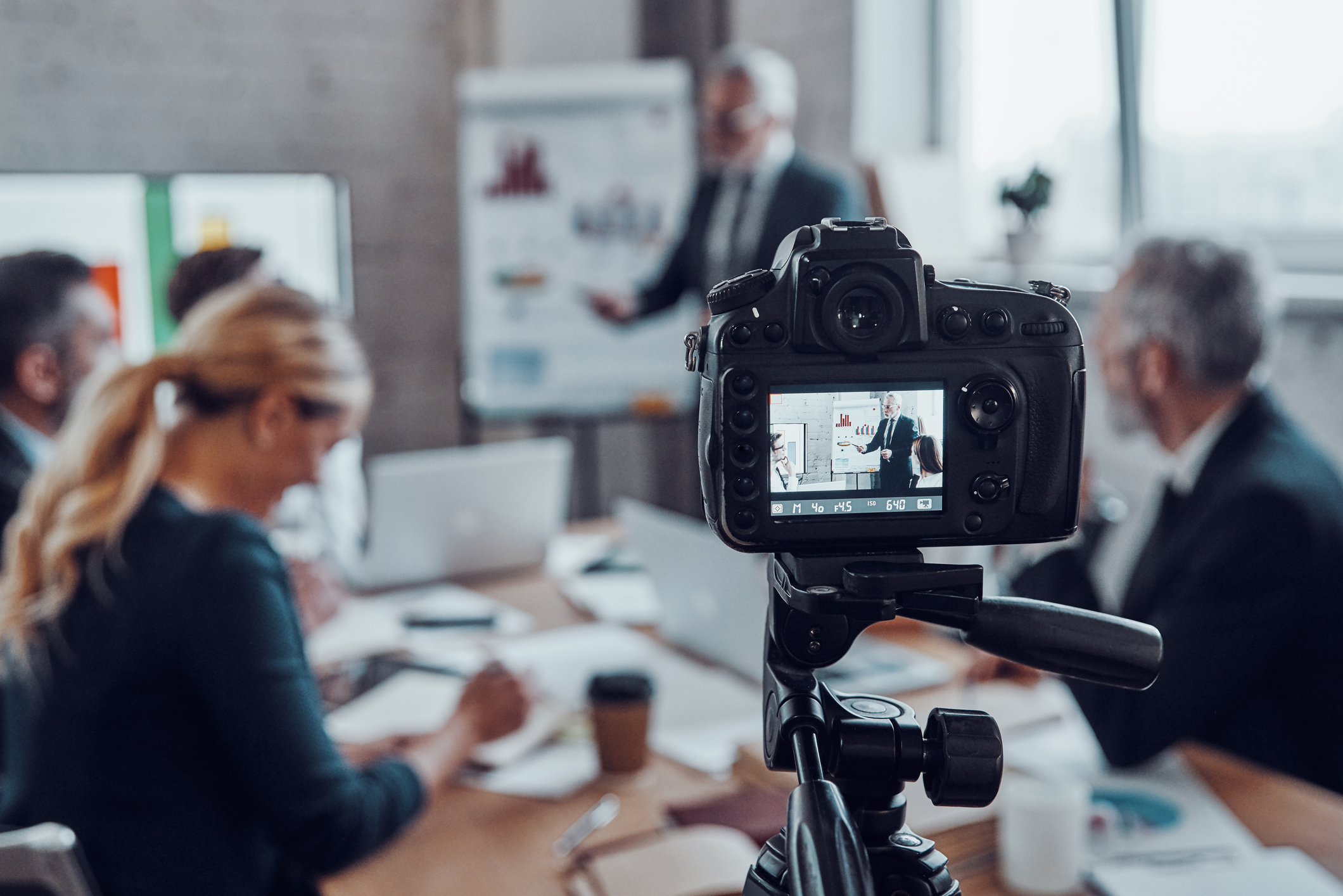 Professional camera on a tripod filming a business meeting where a man in a suit is presenting charts on a whiteboard to seated colleagues.