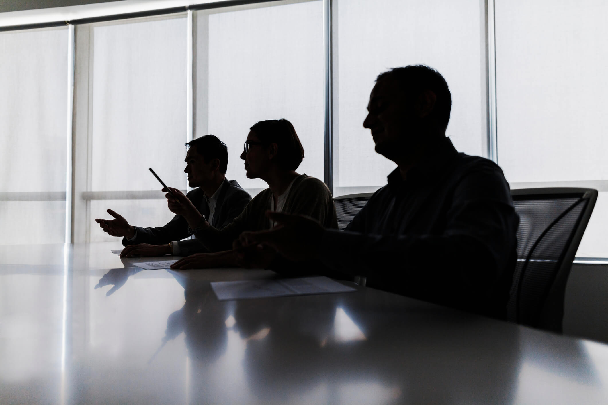 Silhouettes of three people sitting at a conference table engaged in discussion, with a large window and blinds in the background.