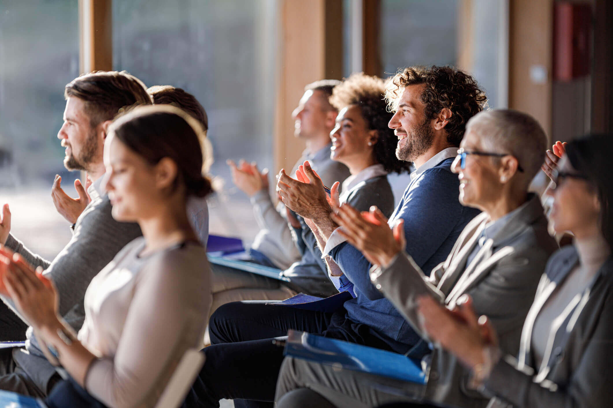 A diverse group of people sitting in a row indoors, smiling and clapping during an event or presentation, with sunlight streaming through windows behind them.
