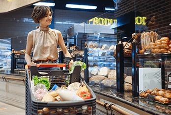 Woman pushing shopping cart filled with fresh vegetables and groceries in a bakery section of a store, looking at the bread on display.
