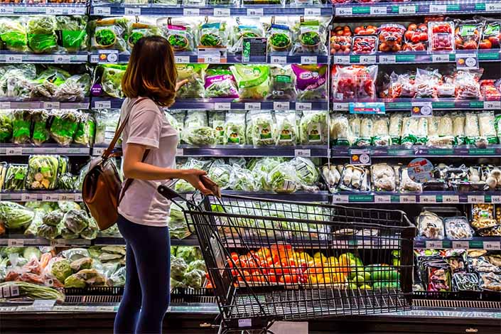 Woman with a brown backpack pushing a shopping cart filled with bell peppers and vegetables, standing in front of refrigerated shelves with various packaged greens and mushrooms in a grocery store.