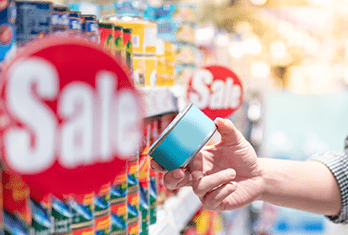 A person holding a canned food item in a store aisle with red Sale signs and various canned goods on shelves.