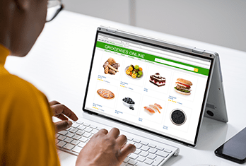 Person using a laptop in tent mode on a white desk, browsing an online grocery store webpage showing various food items like ginger, salad, cake, pizza, blueberries, shrimp, hamburger, and Oreo cookies.