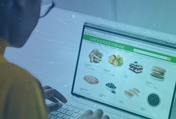 Person browsing an online grocery store on a laptop showing various food items including fruits, baked goods, and snacks available for purchase.