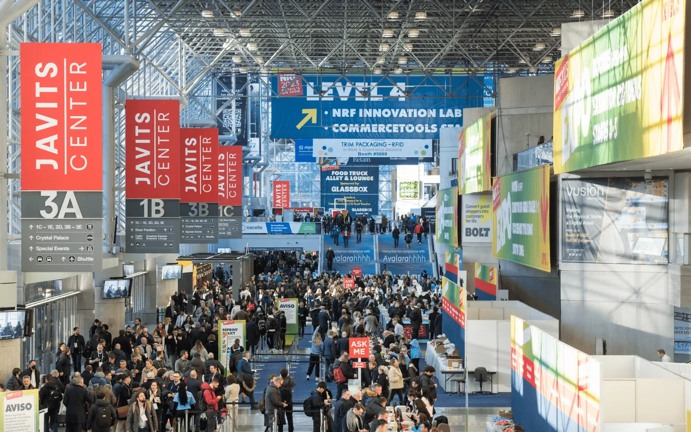 Interior view of Javits Center filled with a large crowd attending an event, with overhead signs indicating sections and booths such as NRF Innovation Lab, CommerceTools, and various sponsors.