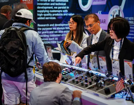 Four people around a display table with electronic equipment and control panels at a trade show booth, with two women and one man behind the table engaging with two visitors, one wearing a white cap and backpack.