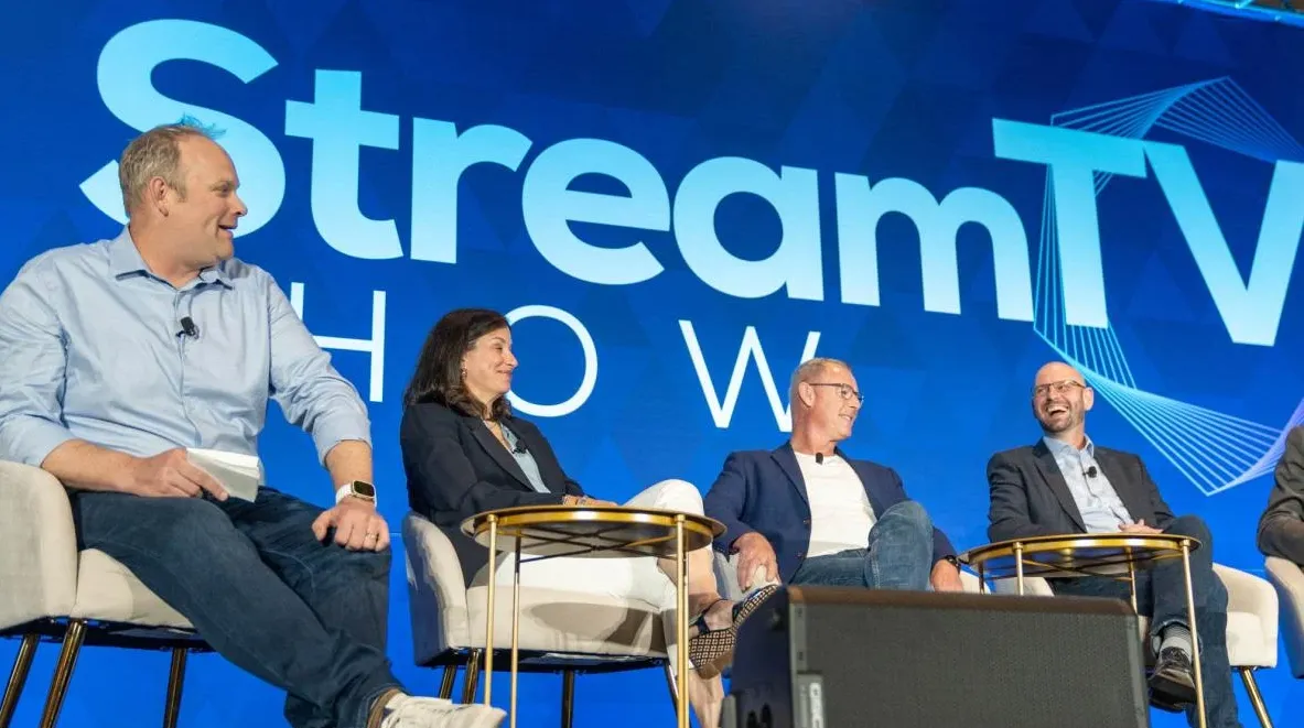 Four panelists sitting on stage in armchairs having a discussion under a large blue screen displaying the text 'Stream TV Show'.