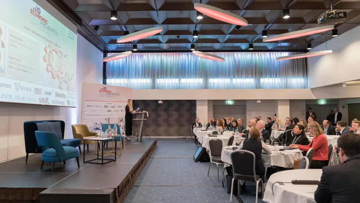 A conference room with attendees seated at round tables facing a stage where a woman is speaking at a podium; a large screen and banner display 'Economic Crime Prevention Switzerland' along with speaker names and logos of sponsors.