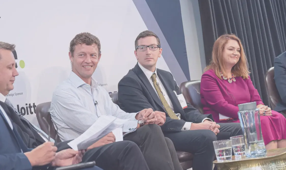 Four professionals seated indoors on stage during a panel discussion, three men and one woman, the woman wearing a purple dress, a table with a glass pitcher and glasses in front of them.