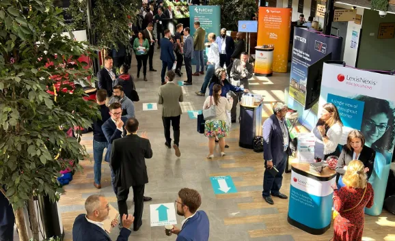 Indoor business event with people walking and talking near exhibition booths and promotional banners for companies including LexisNexis, FullCert, and Sygnia, under natural and artificial light with indoor plants.