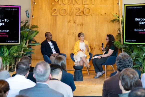 Three panelists seated on a stage with a wooden backdrop displaying 'MONEY 20/20' in large letters, engaged in a discussion before an audience; two screens flank the stage showing event and speaker information.