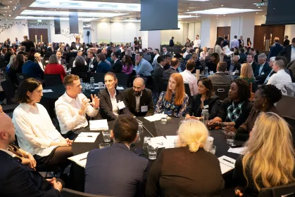 Large group of people seated at round tables in a conference room, engaged in discussion and note-taking during a professional event.