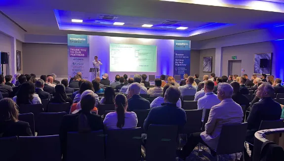 Audience seated in a conference room watching a speaker presenting on stage with projection screen displaying content, flanked by banners reading 'transform finance' and event details.
