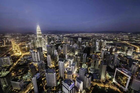 A nighttime aerial view of a cityscape with illuminated skyscrapers, including the brightly lit Petronas Twin Towers, and streets glowing with city lights under a dark blue sky.
