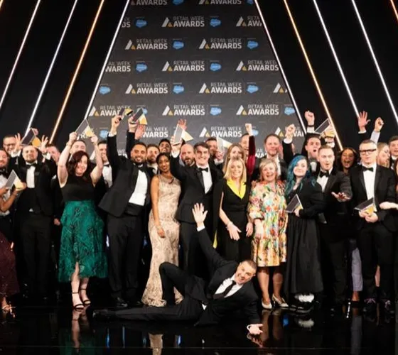 Group of formally dressed people celebrating on stage at the Retail Week Awards, holding trophies and smiling, with a man lying cheerfully in front.