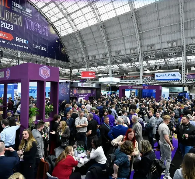 Crowded indoor exhibition hall during a retail technology show with many people interacting near booths and displays under a large arched glass roof.