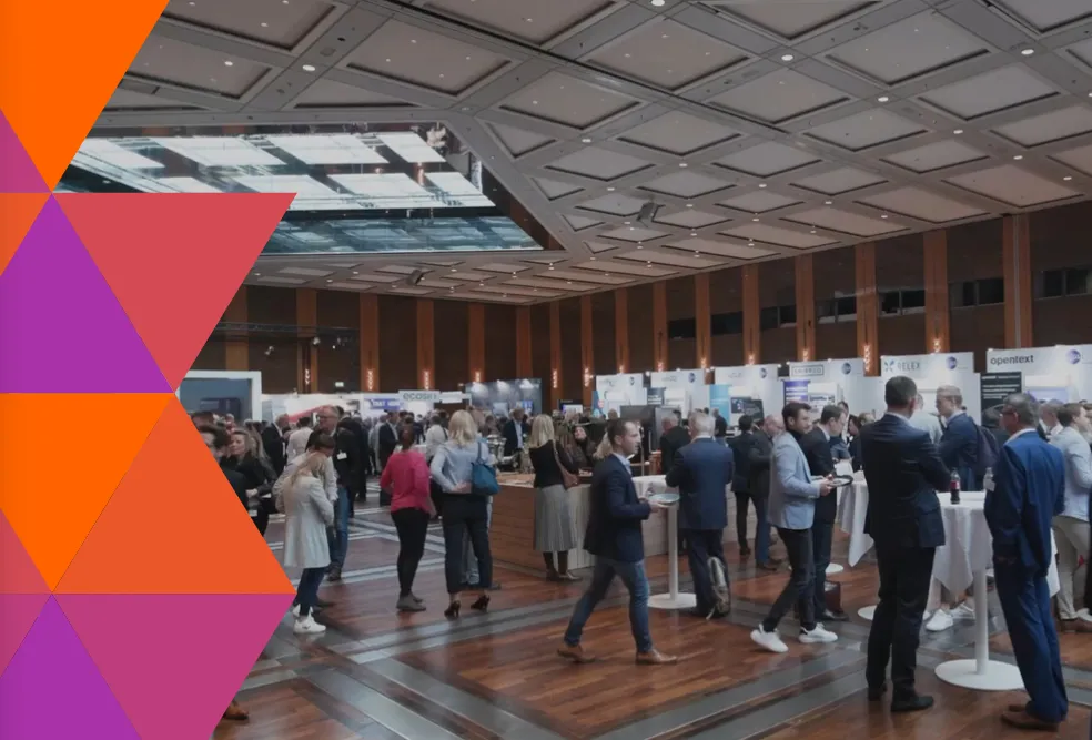 A professional networking event or conference in a large hall with wooden floors and a modern ceiling. People dressed in business attire are standing, walking, and conversing around high tables and exhibition booths with company banners and displays.