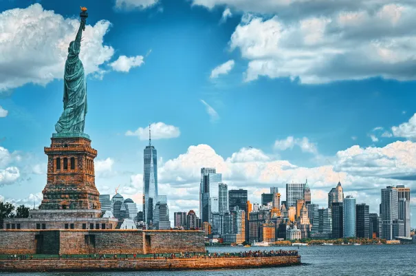 Statue of Liberty on Liberty Island with New York City skyline in the background under a blue sky with clouds.